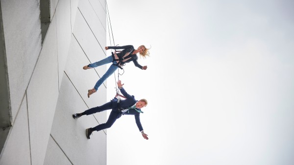 Two people run down a skyscraper wall secured by ropes