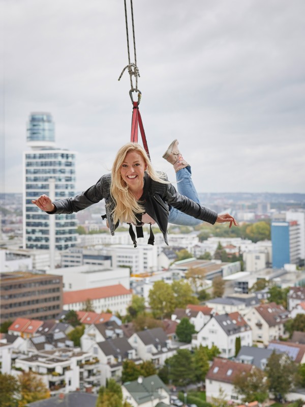 A woman laughing hangs secured on a rope in front of Frankfurt's city panorama