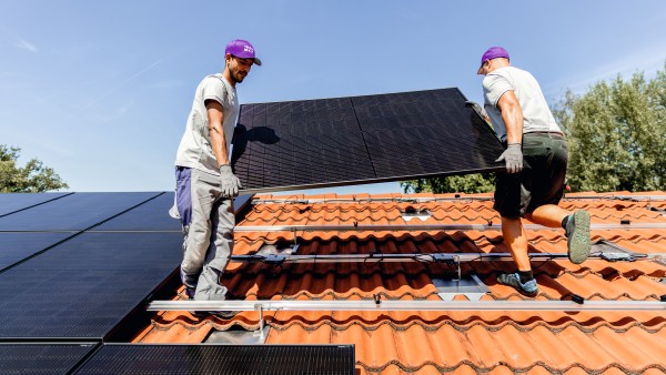 Two installers lift a PV module onto the roof of a house