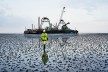 A worker in work clothes stands in the mudflats, behind which one sees a laying ship.