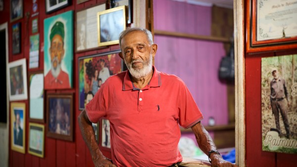 Portrait of Raimundo Mendes Barros, rubber tapper, environmental activist, and cousin of Chico Mendes, standing in front of a wall with many framed photos, including one of Chico Mendes.