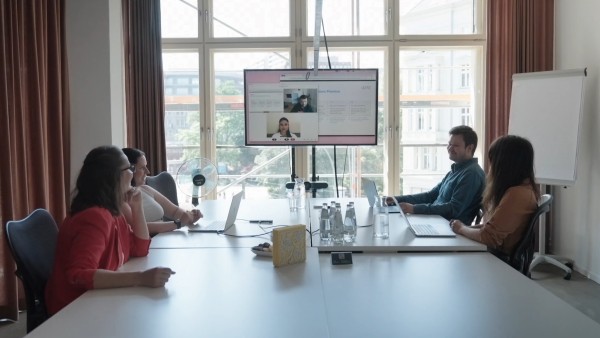 Five people sit around a conference table with laptops and water bottles, a screen shows a video call with two participants.