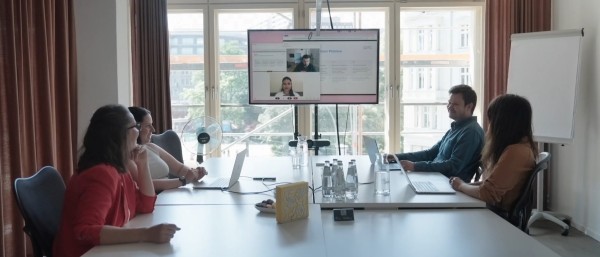 Fünf Personen sitzen an einem Konferenztisch mit Laptops und Wasserflaschen, auf einem Bildschirm ist eine Videokonferenz mit zwei Teilnehmern zu sehen.