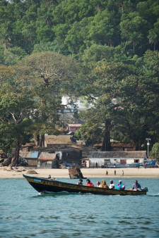 A group of people are transported across the sea on a traditional boat in Guinea. In the background is a beach with residential buildings
