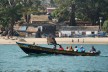 A group of people are transported across the sea on a traditional boat in Guinea. In the background is a beach with residential buildings