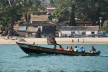 Auf einem tradionellem Boot in Guinea werden eine Gruppr Menschen über das Meer transportiert. Im HIntergrund ist ein Strand mit Wohnhäusern