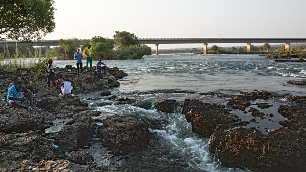 Menschen sitzen auf Steinen am Ufer des Sambesi, im Hintergrund die Brücke
