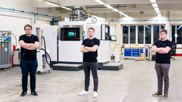 Three men stand with folded arms in front of a large machine. 